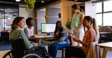 Profissional sorrindo em cadeira de rodas durante reunião em escritório moderno de startup, representando inclusão e cultura de benefícios flexíveis no ambiente de trabalho.
