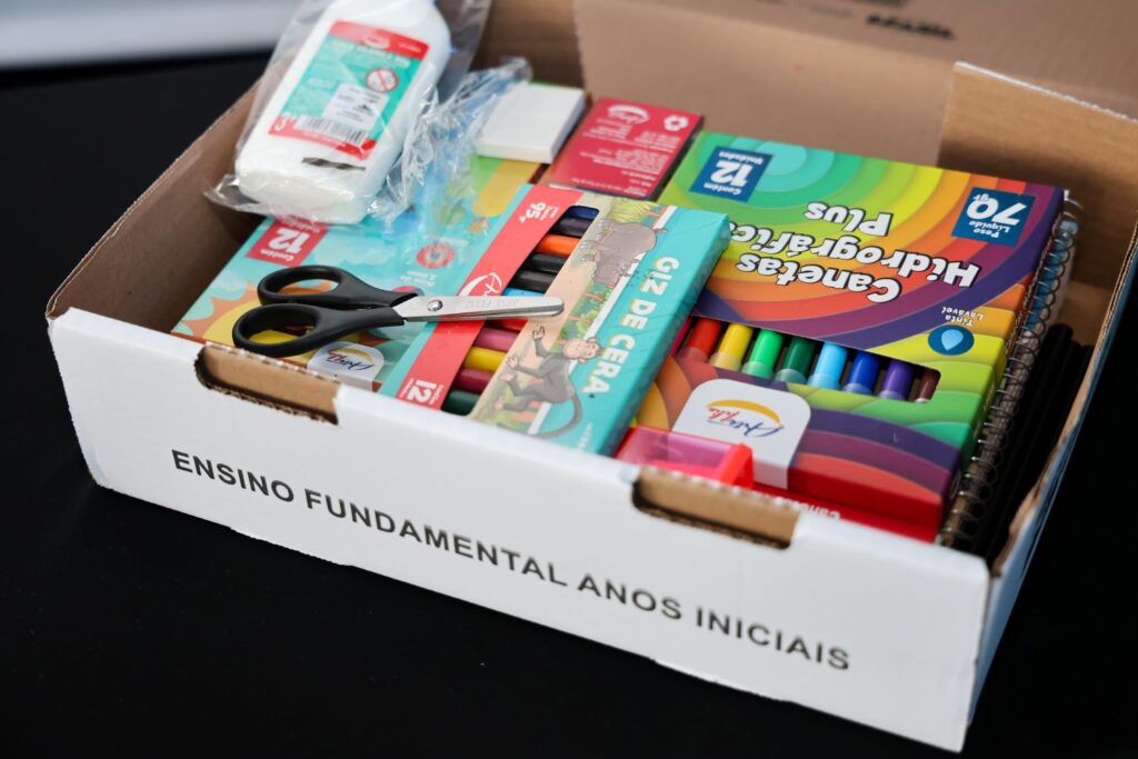 Caixa com kit de material escolar, com cadernos, lápis de cor e tesoura, ilustrando a distribuição física na gestão de benefícios sociais.